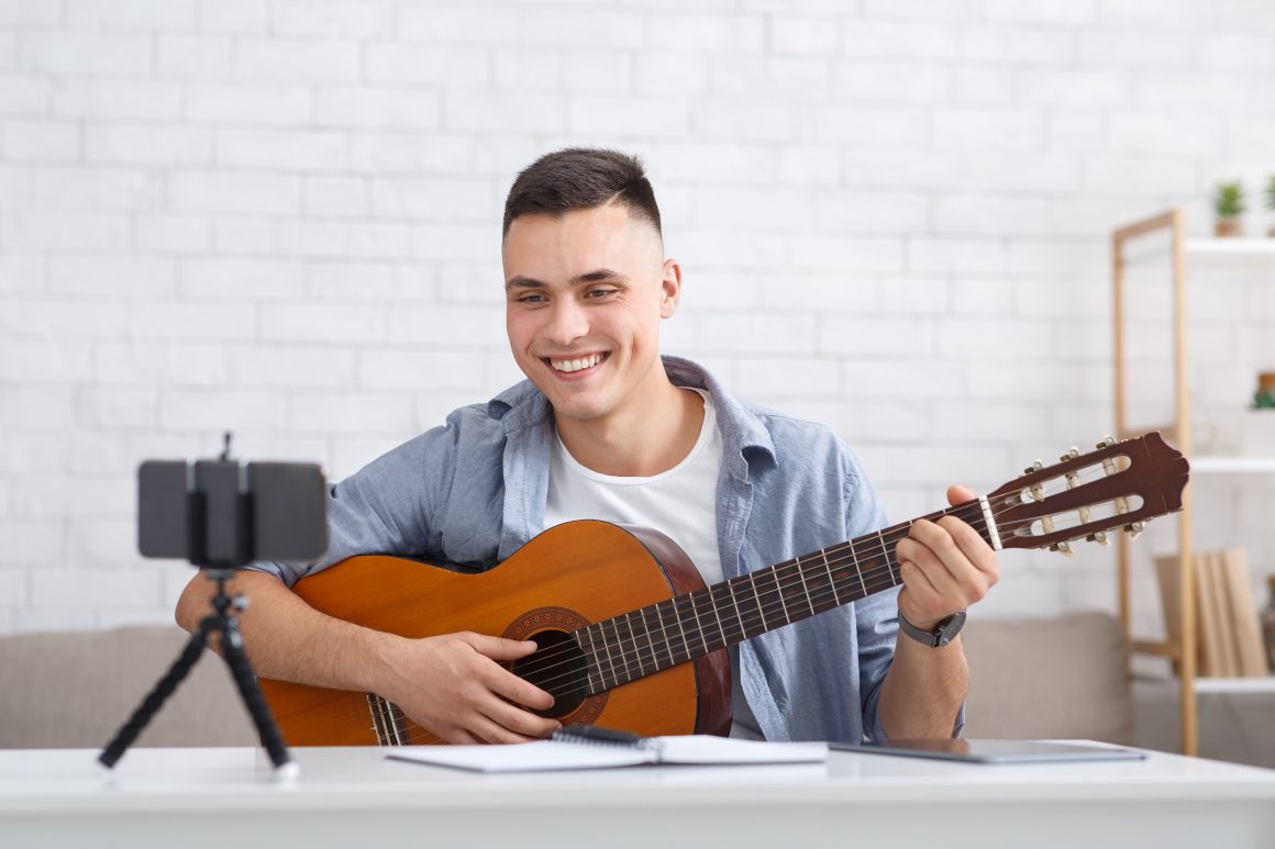 Guitar lesson and online blog. Young happy guy tutor plays on instrument and looks at webcam on smartphone and shoots video for students for study at home Group Classes & Events