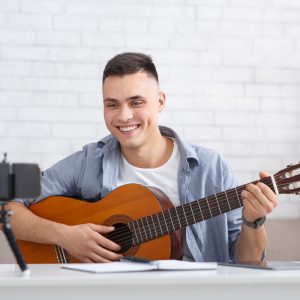 Guitar lesson and online blog. Young happy guy tutor plays on instrument and looks at webcam on smartphone and shoots video for students for study at home Group Classes & Events